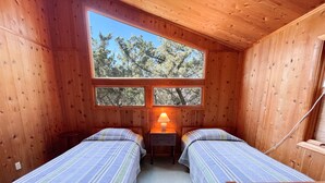 Habitación tipo cabaña con dos camas de madera, una mesita de noche con lámpara y un ventanal que ofrece vistas a árboles.