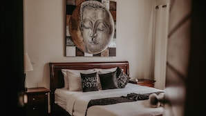 Un lit bien fait, avec du linge de lit blanc et des coussins décoratifs, une tête de lit en bois, une table de chevet avec une lampe, et une œuvre d’art accrochée au mur au-dessus du lit.