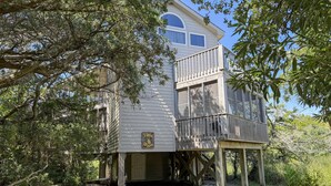 A two-story wooden structure with a balcony, surrounded by dense foliage. - Xenia House:  Enjoy decks and screened porch overlooking the marsh. (Ocracoke)