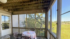 A covered porch with a table and chairs, large windows, and a view of the outdoors. - Xenia House:  Enjoy decks and screened porch overlooking the marsh. (Ocracoke)