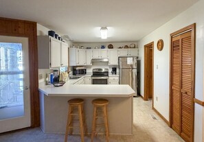 A kitchen with white cabinets, a stainless steel refrigerator, a microwave, and a stove. There are two wooden stools at the kitchen island.