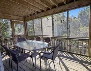 A covered outdoor seating area with a glass-top table and wicker chairs, surrounded by wooden railings and large windows.