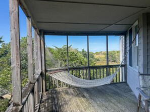 A wooden porch with a hammock, overlooking a landscape with trees and a distant building. - Xenia House:  Enjoy decks and screened porch overlooking the marsh. (Ocracoke)