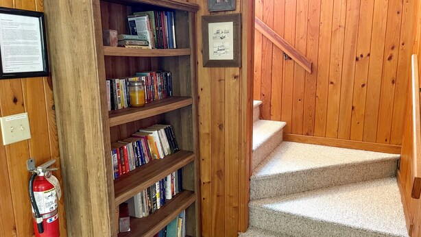 A wooden staircase with a bookshelf filled with books and decorative items. - Xenia House:  Enjoy decks and screened porch overlooking the marsh. (Ocracoke)