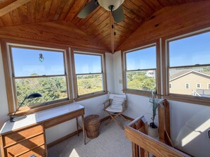 A room with a desk, chair, and large windows offering a view of greenery and buildings. - Xenia House:  Enjoy decks and screened porch overlooking the marsh. (Ocracoke)