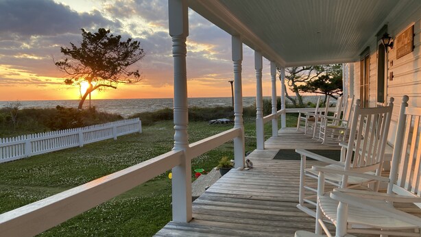 Una terrazza in legno con ringhiere bianche e sedie, affacciata su una spiaggia al tramonto.