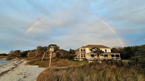 Una casa sulla costa con un ampio portico, circondata da spiaggia e vegetazione, sotto un cielo nuvoloso.