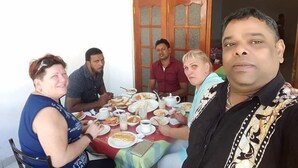 Un groupe de personnes est assis à une table, devant divers plats, et une fenêtre à cadre en bois se trouve à proximité.