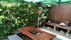 Un área de estar al aire libre con mesas y bancos de madera, rodeada de follaje y una valla de madera.