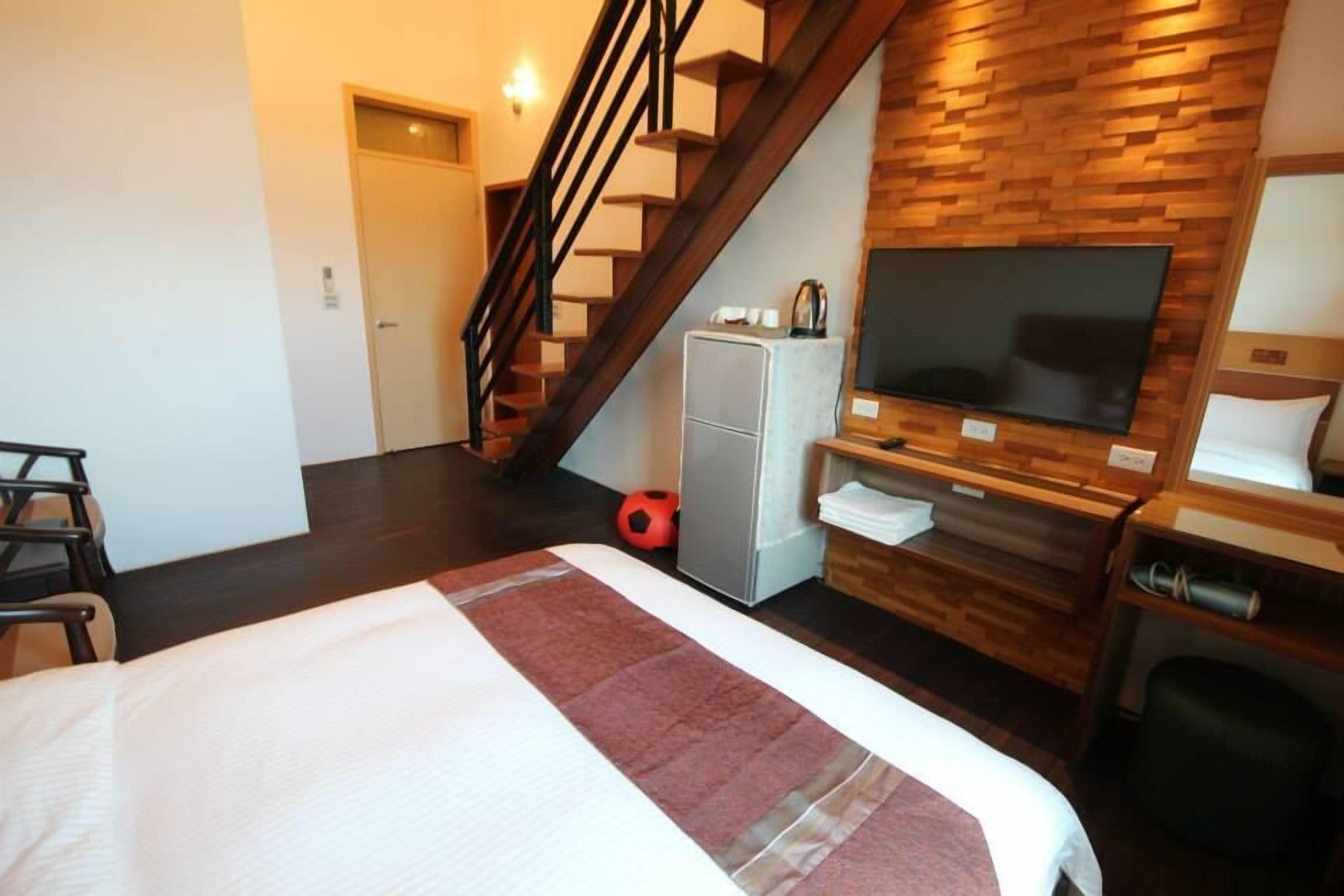Een hotelkamer met een bed, een aan een houten muur bevestigde televisie, een koelkast, een bureau en een trap.