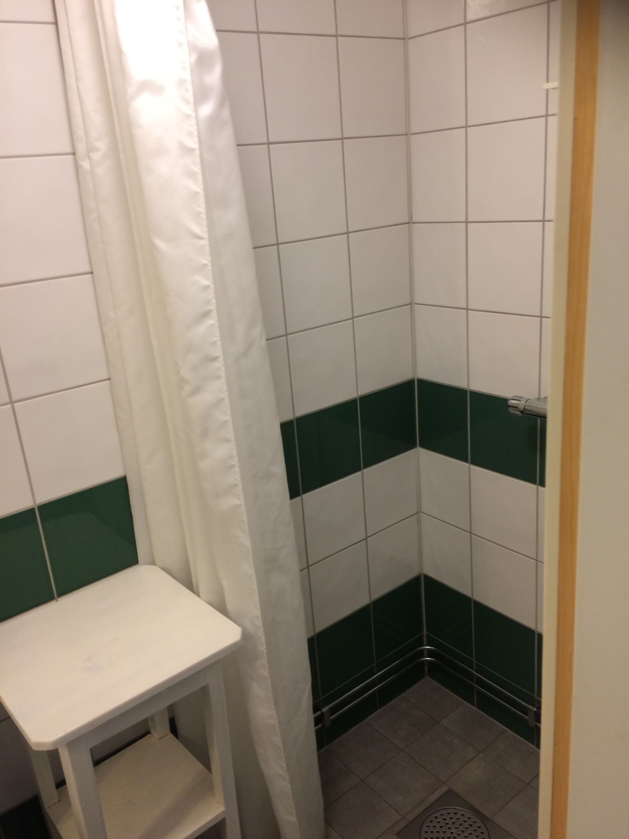 Un baño pequeño con azulejos, un diseño de baldosas blancas y verdes, cortina de ducha blanca y un banco blanco.
