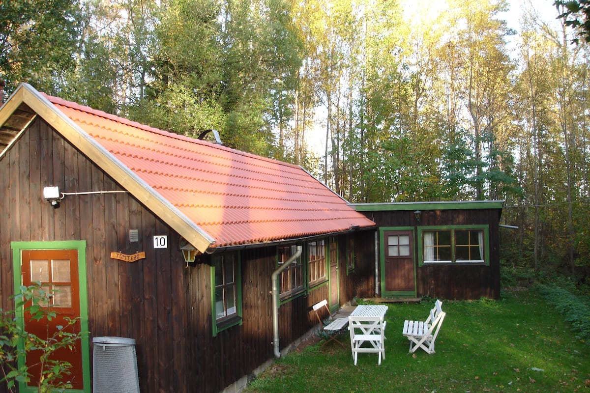 A rustic wooden cabin with a red roof, a green door, and a small attached structure. There is a white table and two chairs outside.
