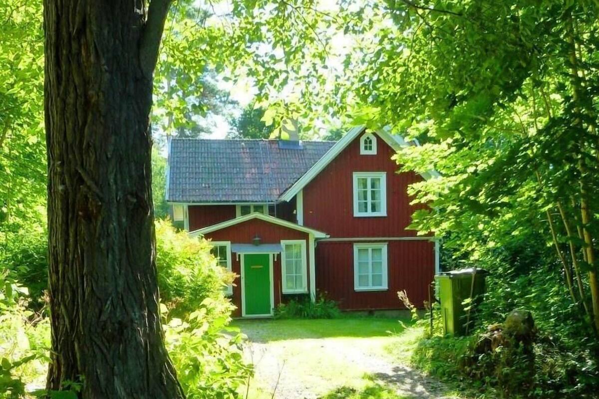 A red house with a green door, surrounded by lush greenery and a large tree in the foreground.