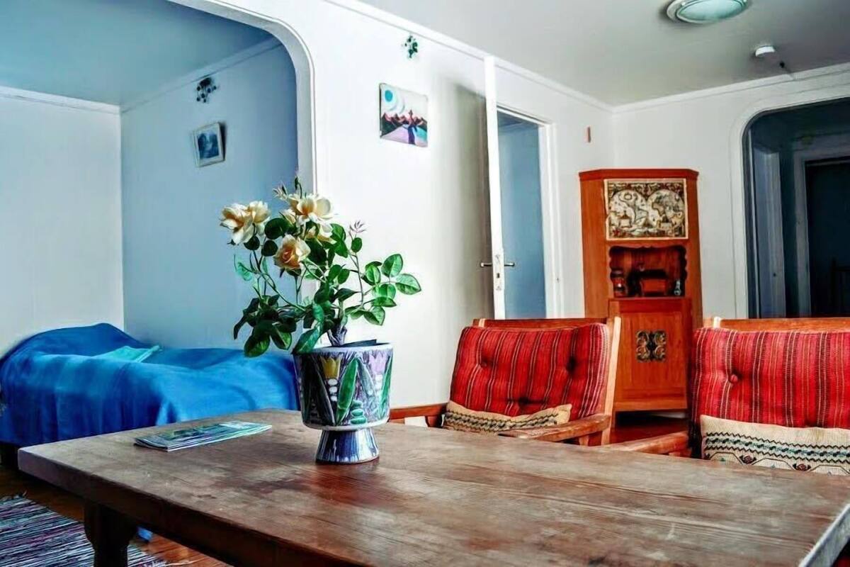 A room with a bed, a wooden table with a vase of flowers, a red upholstered chair, and a wooden cabinet with decorative carvings.