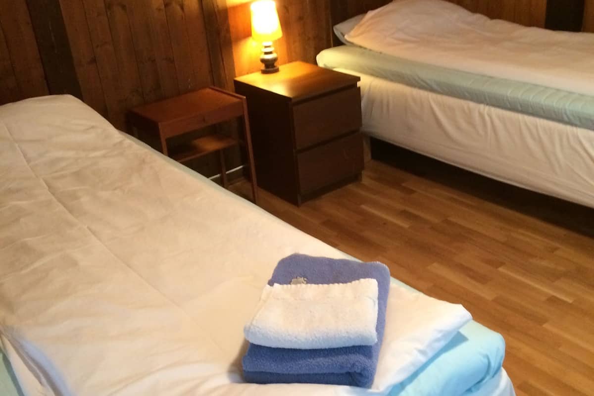 A room with two beds, wooden walls, a nightstand with a lamp, and a small table.