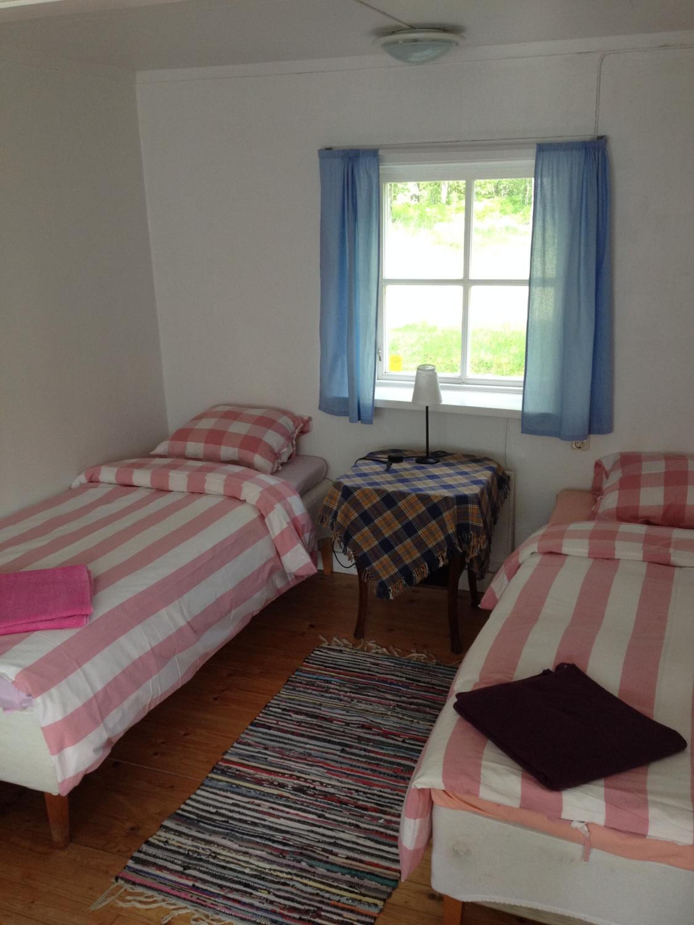A dormitory room with two beds, a small table, a lamp, and a window with curtains.