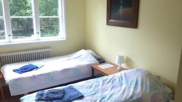 A room with two beds, a window with a view of trees, a radiator, a lamp, and a framed picture on the wall.
