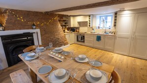 A dining area with a wooden table set for six, a brick wall, and a kitchen in the background. - Charming New Forest Escape Near Forest Walks & Coast (Fordingbridge)