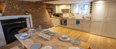 A dining area with a wooden table set for six, a brick wall, and a kitchen in the background.