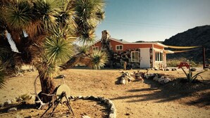 Paisaje desértico con una casa de piedra, una palmera y una pequeña zona de estar al aire libre.