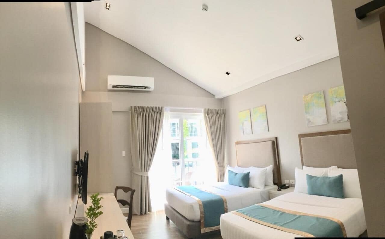 A hotel room with two beds, a TV mounted on the wall, an air conditioning unit, and a window with curtains.