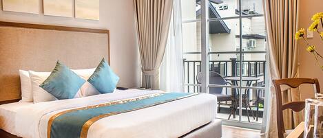 A hotel room with a large bed, a balcony with outdoor seating, and a view of the building outside.