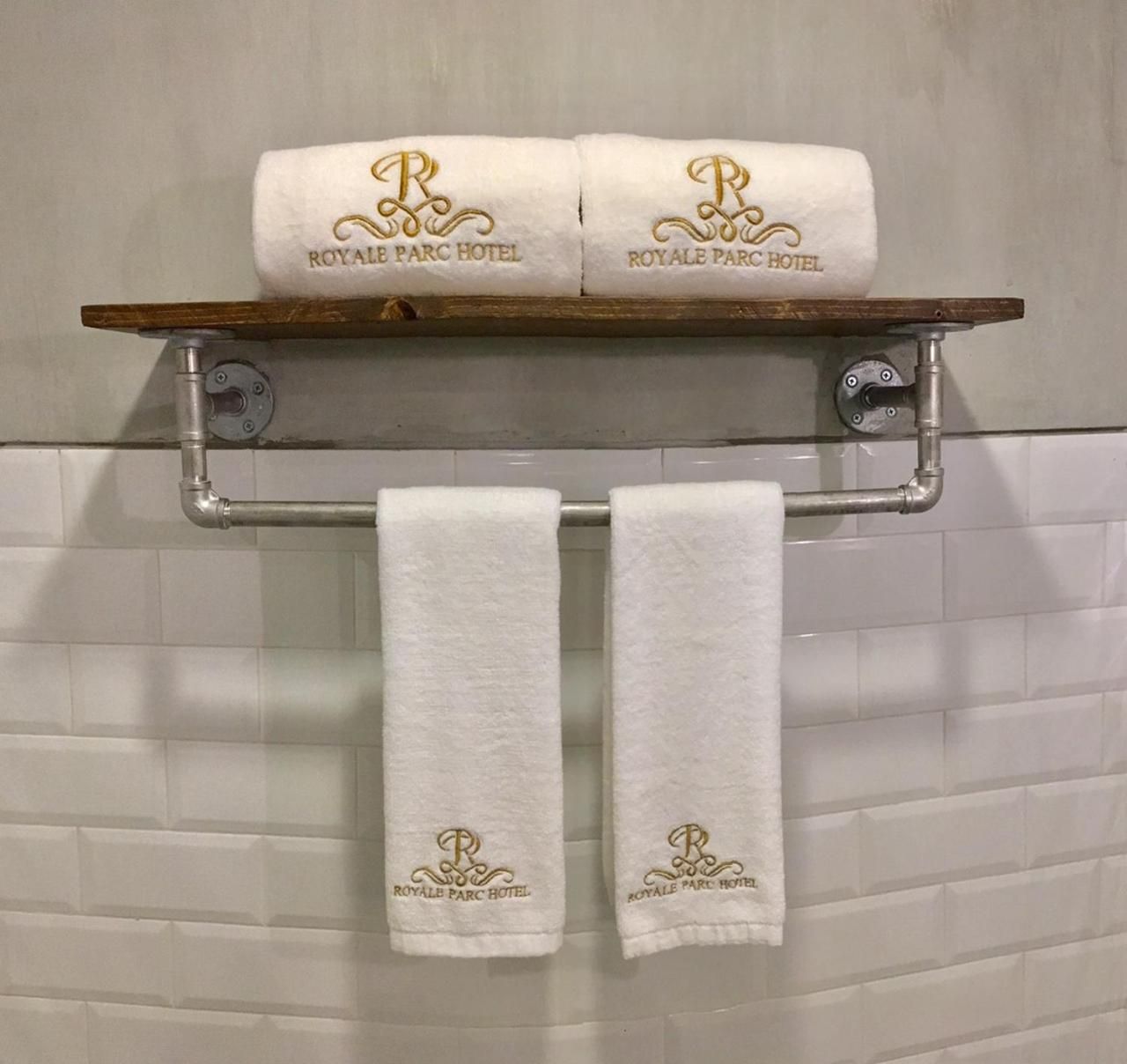 Une salle de bain équipée d’un porte-serviettes où sont posées deux serviettes et d’une étagère sur laquelle reposent deux serviettes enroulées, toutes deux portant le logo « ROYAL PARC HOTEL ».