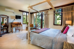 A bedroom with a four-poster bed, a desk with a piano, and large windows offering a view of greenery.