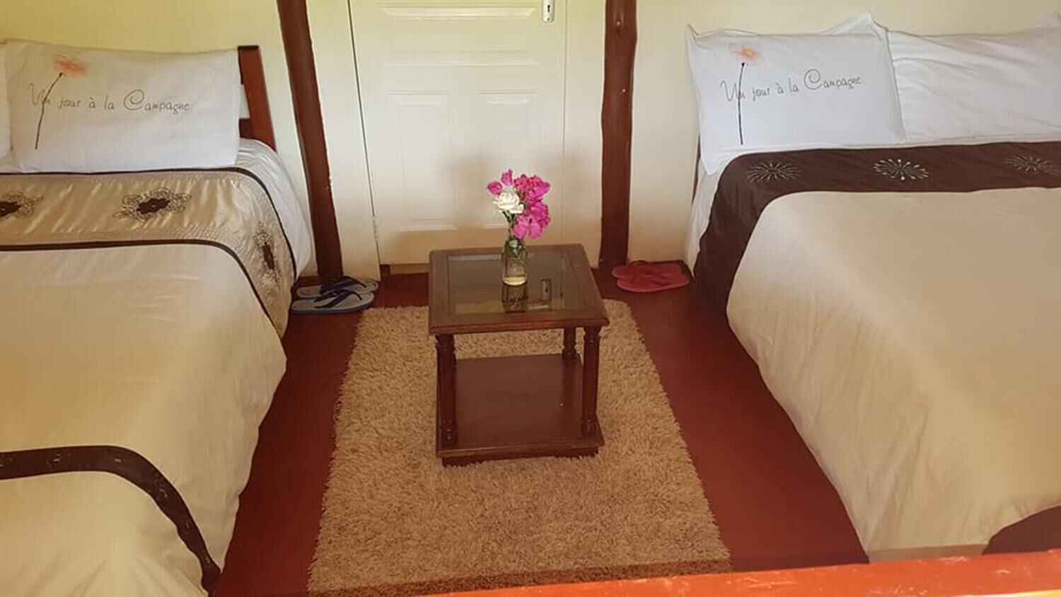A hotel room with two beds, a small table with a vase of flowers, and a door in the background.