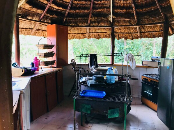 A rustic kitchen with a thatched roof, a gas stove, a sink, and a window overlooking greenery.