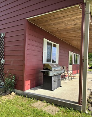 A covered outdoor area with a grill, a wooden deck, and a red building.