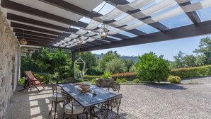 An outdoor patio with a table and chairs, a pergola with a checkered pattern, and a view of greenery and hills in the background.