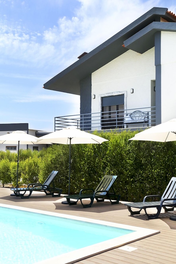 A pool area with lounge chairs and umbrellas, a building with a balcony, and a clear sky.