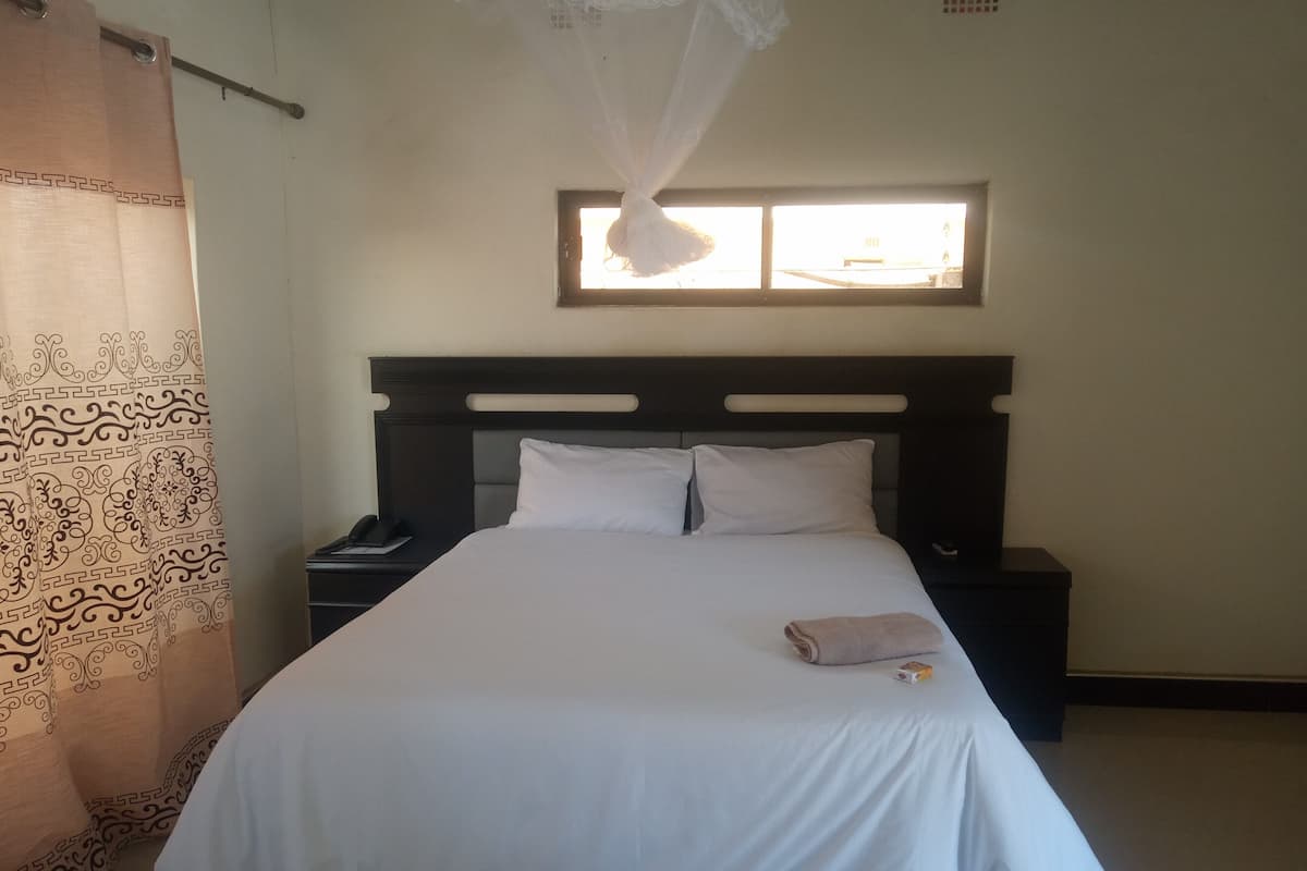 Un lit bien fait, avec du linge de lit blanc, une table de chevet avec un téléphone et une fenêtre donnant sur l’extérieur.