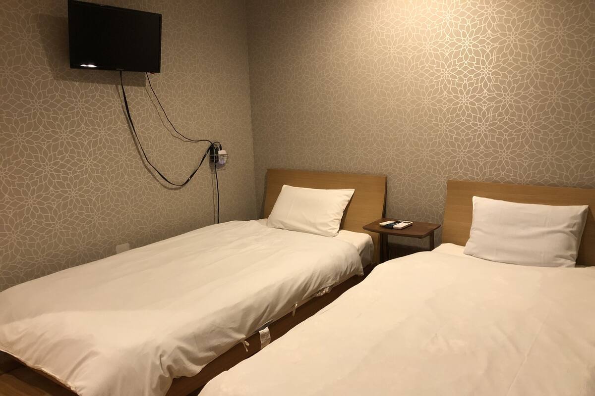 A hotel room with two single beds, a TV mounted on the wall, and a bedside table with a phone and a book.
