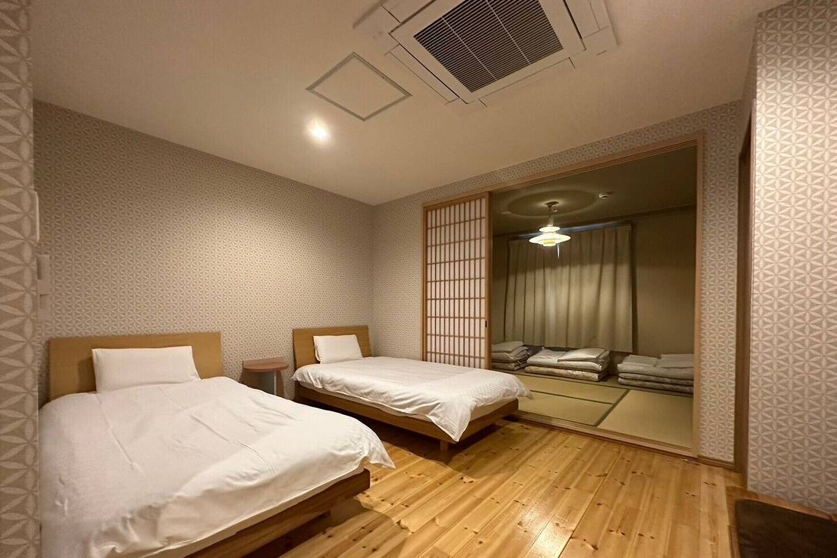 A hotel room with two wooden beds, a small bedside table, a ceiling vent, and a sliding door leading to a bathroom.