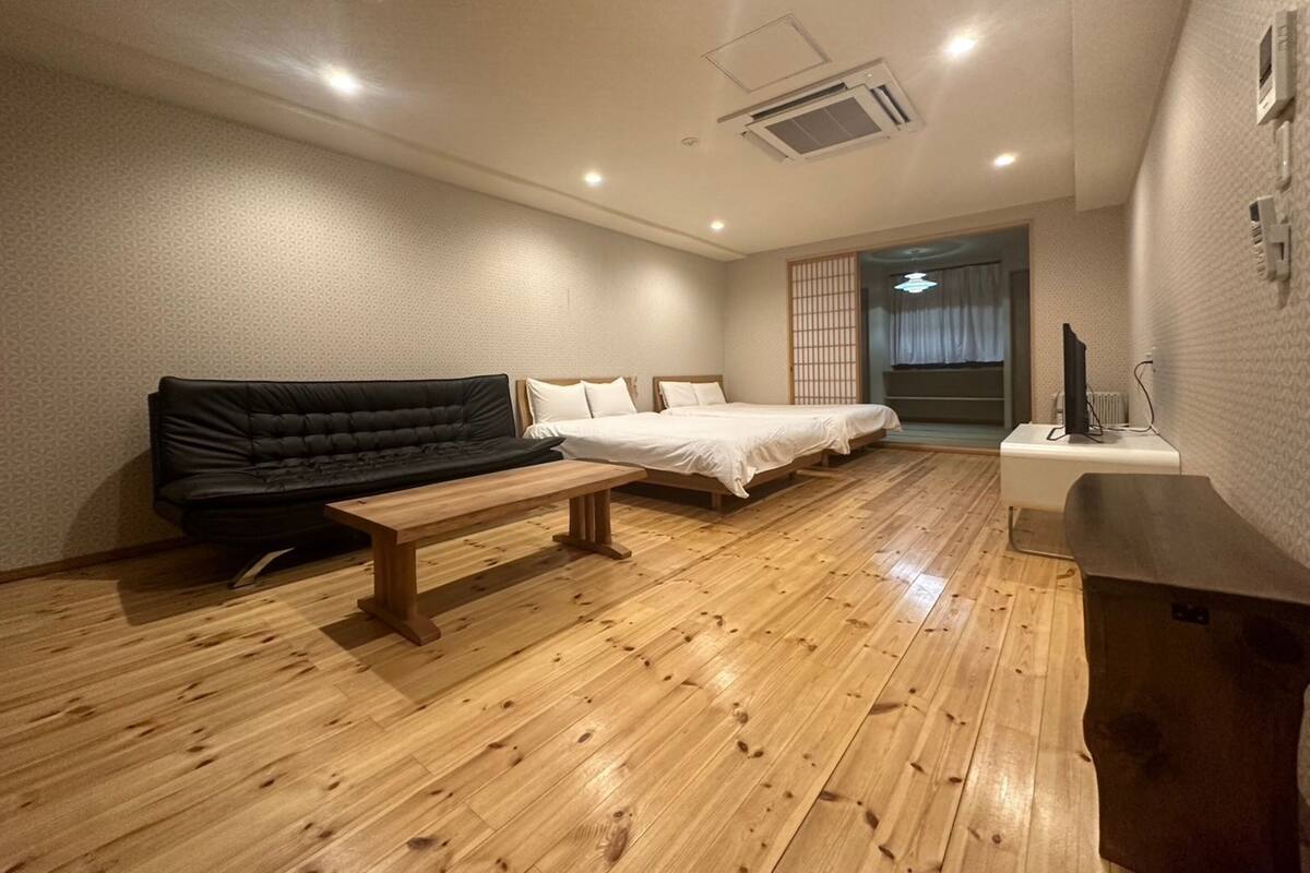 A room with a wooden floor, a black sofa, a bed with white bedding, and a small wooden table.
