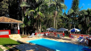 A swimming pool with lounge chairs and umbrellas, surrounded by palm trees and a red fence.