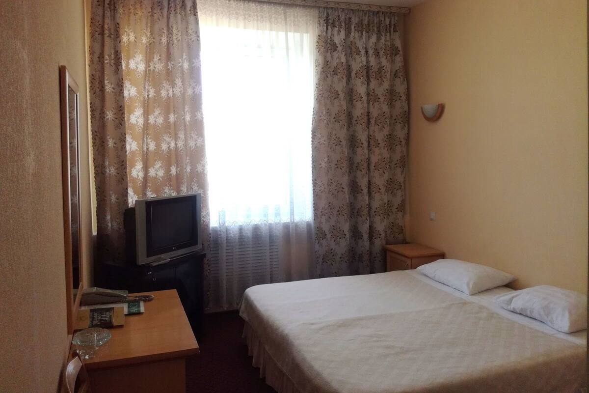 Una habitación de hotel con cama, televisor sobre una mesa de madera y ventana con cortinas.