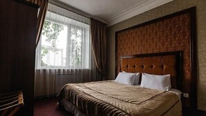 Une chambre d’hôtel avec un grand lit, une fenêtre avec des rideaux, une tête de lit à motifs et une chaise en bois.