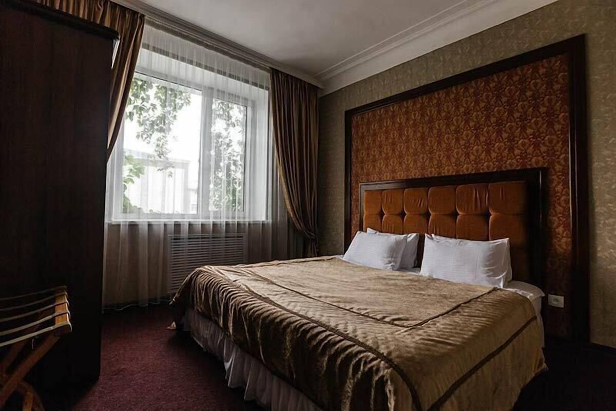 Una habitación de hotel con una cama grande, una ventana con cortinas, un cabecero estampado y una silla de madera.