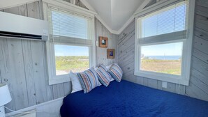 A bedroom with a blue bedspread, two windows with blinds, a wooden wall, and a small picture frame. - Mosby's Manor:  Eclectic design, unique interior, distant sound views. (Ocracoke)