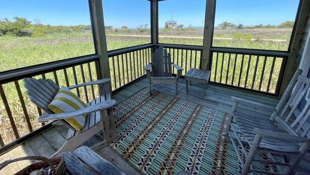 A covered porch with wooden railings, two Adirondack chairs, a small table, and a patterned rug. - Mosby's Manor:  Eclectic design, unique interior, distant sound views. (Ocracoke)