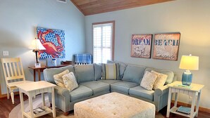 Uma sala de estar com sofá seccional, uma mesinha, uma poltrona e um tapete. Há duas obras de arte nas paredes e um abajur em uma mesa lateral.
