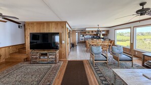 Ein geräumiges Wohnzimmer mit großem Flachbildfernseher, Holzboden, einem Essbereich mit Holzsesseln und einer Küche im Hintergrund.