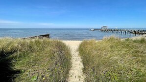 Ein Strandweg führt zu einem hölzernen Steg, der sich ins Meer erstreckt, mit einer kleinen Schutzhütte am Ende.