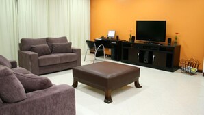 A living room with a brown sofa, a brown ottoman, a black entertainment unit, and a television. - Residence Plaza Flat (Ribeirão Preto)