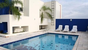 A swimming pool with tiled edges, surrounded by a white building with a blue accent wall and a ladder. - Residence Plaza Flat (Ribeirão Preto)