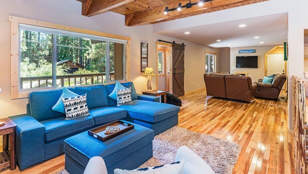 A spacious living area with a blue sectional sofa, wooden flooring, and a large window offering a view of the outdoors.