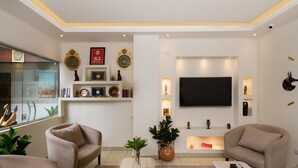 Un salon moderne avec deux fauteuils beiges, une table basse, un téléviseur à écran plat fixé au mur et une bibliothèque agrémentée d’objets décoratifs.